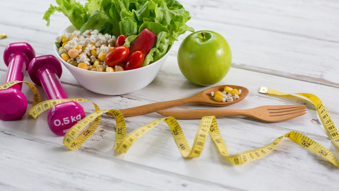 A table with a health bowl of vegetables, an apple, medications, a tape measure and exercise weights