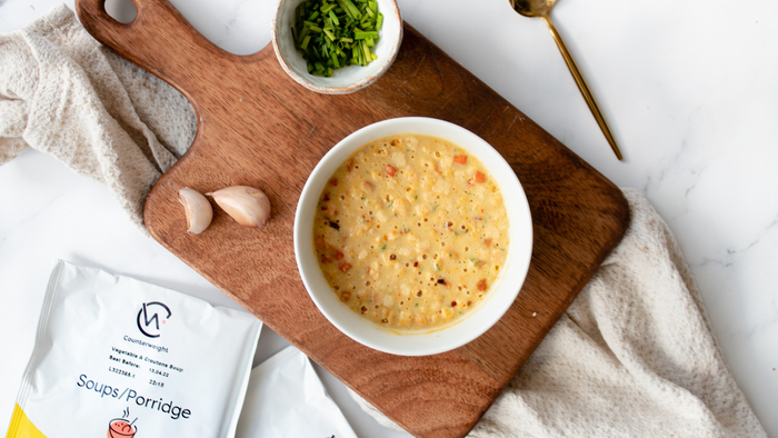Counterweight soup/porridge packets next to a prepared soup with garlic gloves, chives and spoon on a serving board with a cloth