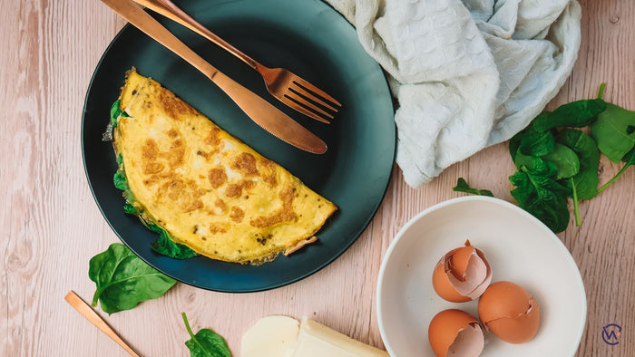 A plate with an omelette with greens, knife and fork next to a bowl of egg shells