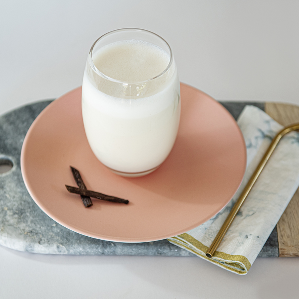 A prepared vanilla shake in a glass on a plate with a gold reusable straw on a napkin on a board with two pieces of vanilla next to the glass. 